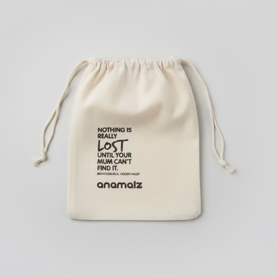 A beige fabric drawstring pouch with black printed text, including the brand name 'anomaliz' and a quote, laid flat on a wooden surface.