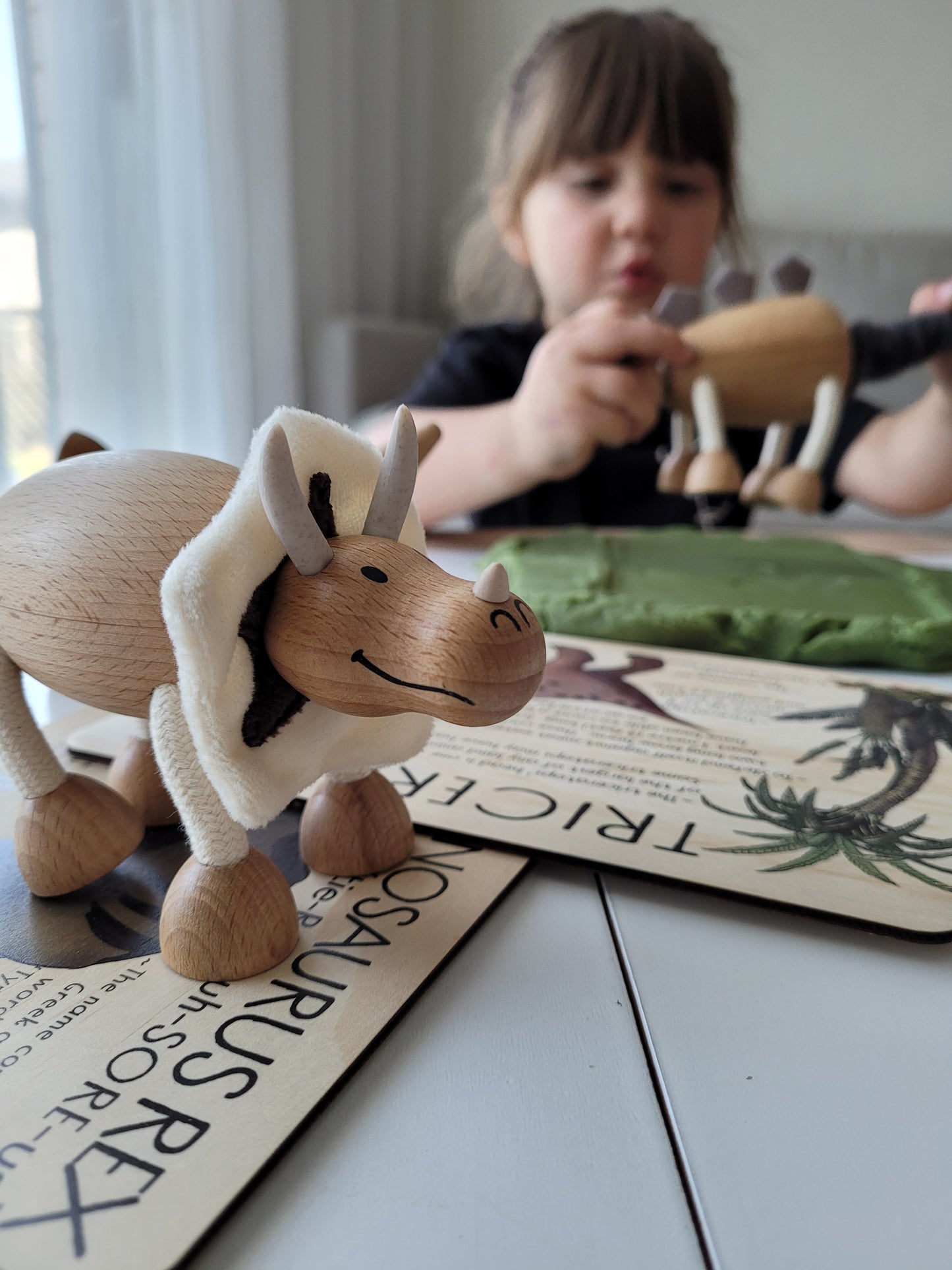 wooden dinosaur toy on a table with a small girl in the background playing another wooden dinosaur toy 